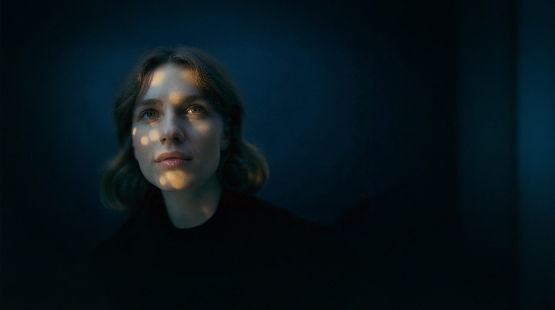 A woman looks up in quiet wonder, warm dappled light falling across her face against deep blue.