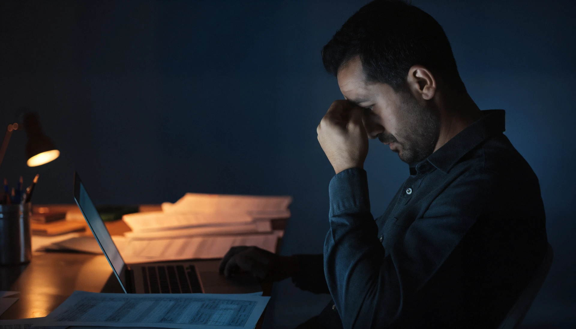 A producer late at night at his desk, head in his hand, a laptop and printed spreadsheets under the lamp.